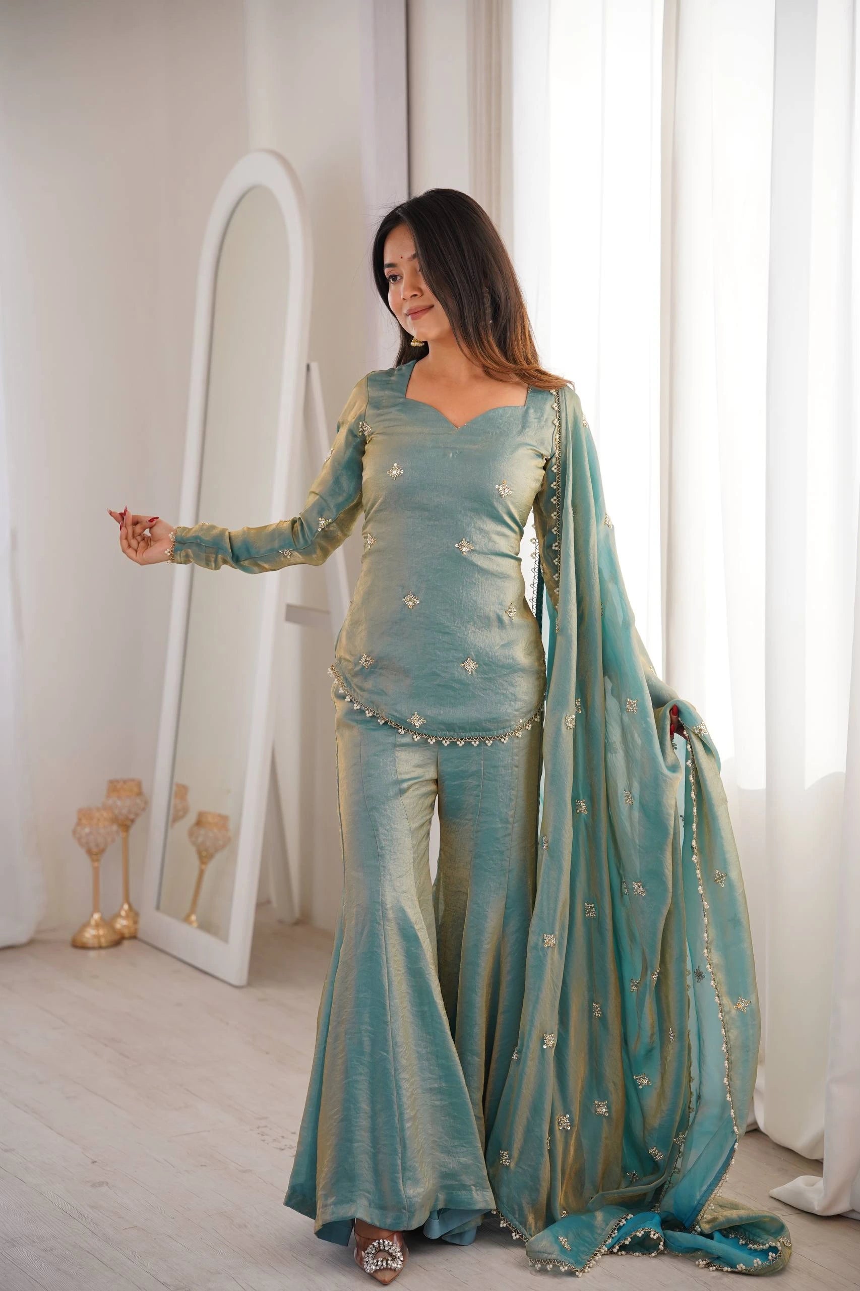 Woman in a teal traditional outfit standing in a room with a mirror and white curtains.